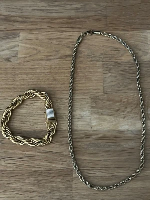  guld armband & halsband  - Säljer ett armband och halsband i guldton med tvinnad kedja och ett rektangulärt spänne dekorerat med små glittrande stenar. Perfekt för dig som gillar statement-smycken och vill sticka ut lite extra.