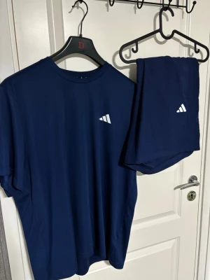 Blått träningsset Adidas - Snyggt blått träningsset från Adidas med t-shirt och shorts. Båda plaggen har den klassiska Adidas-loggan i vitt på bröstet och benet. Tillverkade i ett lätt och snabbtorkande material som passar perfekt för träning och sport.