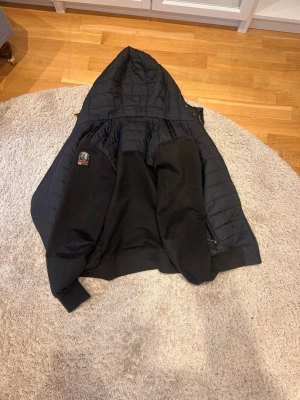 Svart vårjacka från Parajumpers - Säljer en svart pufferjacka från Parajumpers med huva och quiltad design. Jackan har en tydlig logotyp på bröstet och är tillverkad i ett slitstarkt material som håller värmen under kalla dagar. Perfekt för dig som vill ha en snygg och funktionell vårrjacka.