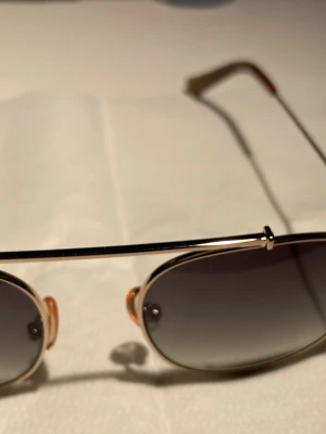 Guldiga aviator solglasögon Vasuma by EWear DESIGN OF SWEDEN  - Stilrena aviator solglasögon med tunn guldfärgad metallbåge och mörktonade linser. Klassisk form med näskuddar och smala skalmar. Perfekta för soliga dagar och ger en tidlös vibe till din outfit.