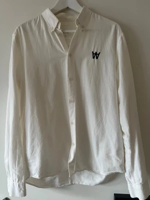 Vit skjorta från Wood Wood - Klassisk vit skjorta från Wood Wood med svart broderad W-logga på bröstet. I bra skick, klassisk och stilren!