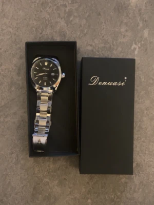 Silverfärgad klocka från Denuasi - Stilren klocka från Denuasi med svart urtavla och silverfärgad länk i metall. Klassisk rund form och tydliga markeringar på urtavlan. Perfekt accessoar för dig som gillar en clean och tidlös look.