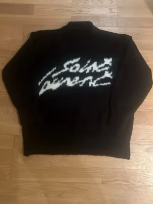 Svart stickad Saint Laurent tröja - Cool svart stickad tröja från Saint Laurent med vit grafisk text på bröstet. Oversized passform och rund halsringning. Perfekt för dig som gillar streetwear och vill ha något unikt i garderoben.