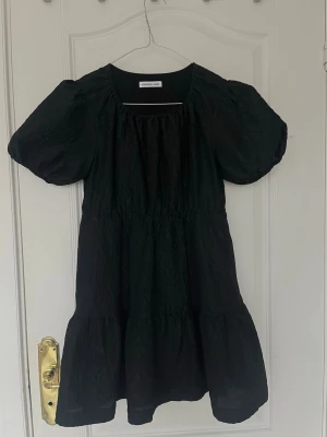 Svart kort klänning Designers Remix - Supersöt svart kort klänning från Designers Remix med puffärmar och lätt rynkad struktur. Klänningen har rund halsringning och en luftig passform som ger en avslappnad men ändå trendig look. Perfekt för dig som gillar stilrena och moderna plagg.