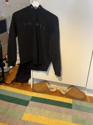 Svart hoodie från Röhnisch - Säljer en svart hoodie från Röhnisch med diskret logga framtill. Hoodien har huva med dragsko och ribbade muddar vid ärmslut och nederkant. Perfekt för en avslappnad och sportig stil.