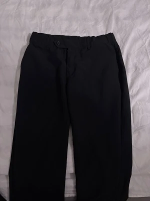 Svarta kostymbyxor med rak passform - Snygga svarta kostymbyxor med rak passform och klassisk midja. Byxorna har knapp och gylf framtill samt hällor för skärp. Perfekta för en stilren look. Materialet ser ut att vara en mjuk och slät textil. Low waist