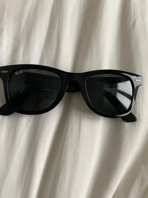 Svarta Ray-Ban Wayfarer solglasögon - Klassiska svarta Wayfarer solglasögon från Ray-Ban med rektangulära bågar och mörka glas. Snygg och tidlös design med Ray-Ban-logga på sidan. Perfekta för att lyfta din stil under soliga dagar.