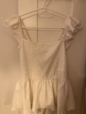 Vit volangblus med knyt i ryggen - Supersöt vit blus med volangdetaljer på axlarna och knytning i ryggen. Blusen har broderade hålmönster och en luftig, flowy passform. Perfekt för varma dagar och ger en romantisk vibe till din outfit.