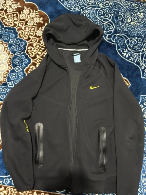 Svart Nike NOCTA hoodie med gul logga - Säljer en svart hoodie från Nike NOCTA-kollektionen med gul Swoosh på bröstet och NOCTA-tryck på ärmen. Hoodien har dragkedja framtill, två fickor med svarta detaljer och huva. Små gula tryck på ryggen ger extra stilpoäng. Perfekt för streetwear och chill.
