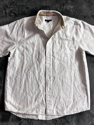 Vit/beige skjorta från Burberry  - Klassisk vit skjorta från Burberry med korta ärmar och bröstficka. Skjortan har knappar framtill och en snygg detalj med det klassiska rutiga mönster på insidan av kragen. Perfekt till sommaren och enkel att matcha. Storlek medium men passar L 