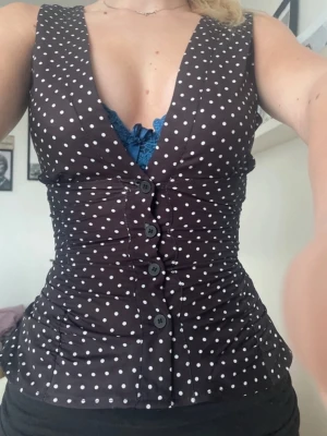 Svart prickig blus med knappar - Snygg svart blus med vita prickar och djup v-ringning. Toppen har knappar framtill och är ärmlös