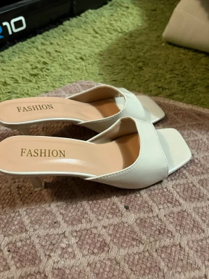 Vita slip-in pumps med låg klack - Stilrena vita slip-in pumps från Fashion med öppen häl och fyrkantig tå. Skorna har en låg klack och bred rem över foten. Tillverkade i ett slätt, glansigt material som ger en modern look. Perfekta för dig som gillar minimalistisk och trendig stil.