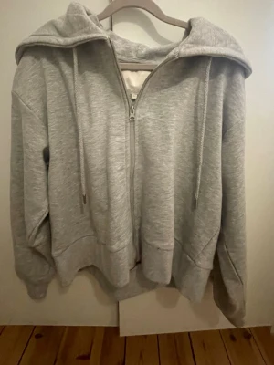 Grå zip hoodie med huva - Säljer en grå hoodie med dragkedja framtill och stor huva. Hoodien har långa ärmar, ribbade muddar och snören i huvan. Perfekt för en avslappnad och chill stil. Materialet känns mjukt och bekvämt, troligen bomullsblandning.