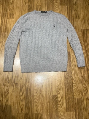 Ljusgrå kabelstickad tröja Polo Ralph Lauren - Snygg ljusgrå kabelstickad tröja från Polo Ralph Lauren med klassisk rund halsringning och den ikoniska broderade loggan på bröstet. Tröjan har långa ärmar och ribbade muddar vid ärmslut och nederkant. Perfekt för lager på lager och stilrena outfits.