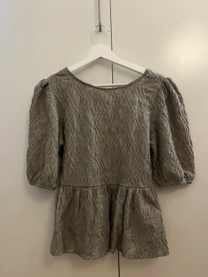 Beige strukturerad blus med knyt - Supersöt beige blus med strukturmönster och puffiga trekvartsärmar. Blusen har en vid ringning och en snygg knytning i ryggen. Tillverkad i ett lätt, krinklat tyg som ger en avslappnad men trendig look.