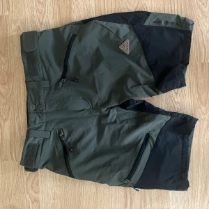 Gröna och svarta shorts från Tenson - Säljer ett par gröna och svarta shorts från Tenson med flera dragkedjeförsedda fickor och justerbar midja med kardborre. Shortsen har en sportig look och är perfekta för utomhusaktiviteter. Materialet känns slitstarkt och funktionellt.