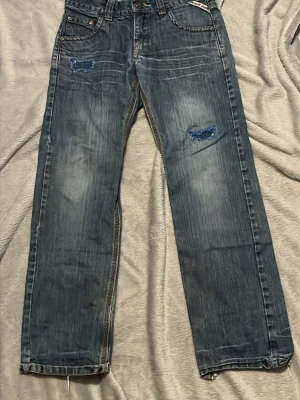 Blå raka jeans från Detroit denim - Säljer ett par blå raka jeans från Detroit denim med slitningar och patchdetaljer framtill. Jeansen har klassiska fem fickor, dragkedja vid benslut och en stor logopatch bak i midjan. Materialet är denim i bomull med snygg tvätt och kontrastsömmar.