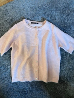 Ljusrosa kofta från 157 med pärlknappar - Supersöt ljusrosa kofta från 157 med korta ärmar och rund halsringning. Koftan har pärlknappar framtill och är stickad i ett mjukt material som känns fluffigt. Perfekt att slänga över en topp för en söt och enkel look.