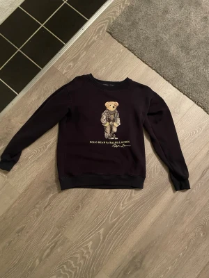 Mörkblå sweatshirt Polo Bear Ralph Lauren - Cool svart sweatshirt från Ralph Lauren med den ikoniska Polo Bear-trycket på bröstet. Tröjan har rund halsringning, långa ärmar och ribbade muddar. Perfekt för dig som gillar streetwear och vill ha något klassiskt men ändå lekfullt.