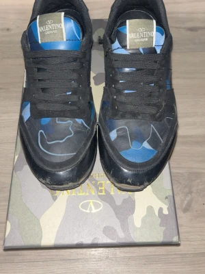 Valentino Garavani blå camo sneakers - Snygga sneakers från Valentino Garavani med blå och svarta camo-detaljer, svarta skosnören och svart sula. Ovandelen är i en mix av mocka och textil med coola mönster och logga på plösen. Perfekta för dig som gillar streetstyle och vill sticka ut.