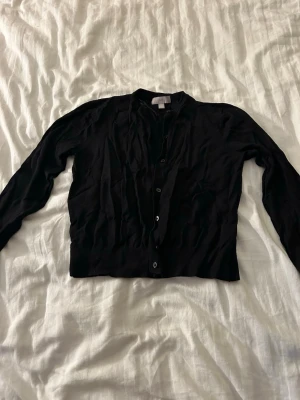 Svart kofta från H&M, storlek M - Klassisk svart kofta från H&M med rund halsringning och knäppning framtill. Tillverkad i ett mjukt och tunt material som passar perfekt för lager på lager. Långa ärmar och ribbade muddar vid ärmslut och nederkant.