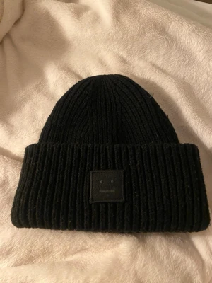 Acne mössa  - Säljer en svart ribbstickad mössa från Acne Studios med den klassiska logotypen framtill. Mössan har bred uppvikt kant och är tillverkad i mjukt material som håller dig varm.