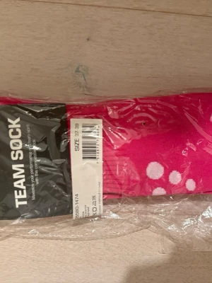 Rosa Team Sock med prickar - Säljer ett par sportiga rosa Team Sock-strumpor med vita prickar. Strumporna är tillverkade i ett syntetmaterial som passar perfekt till träning. De har en klassisk form och är designade för att ge bra stöd under aktivitet. Helt nya.