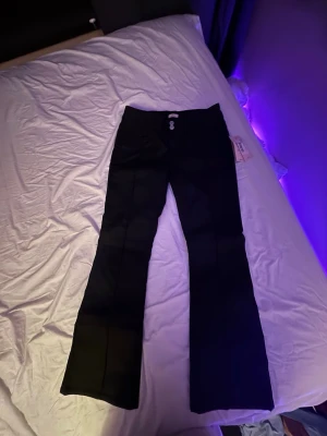Svarta bootcut skinnbyxor  - Säljer ett par svarta skinnbyxor med bootcut passform och pressveck. Byxorna har två knappar framtill och är tillverkade i ett slätt tyg som ger en clean look. Perfekta för dig som vill ha en stilren och trendig siluett.