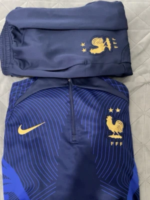 Frankrike Nike träningsset blå/gul - Snyggt träningsset från Nike för Frankrikes landslag, bestående av en långärmad tröja och byxor i mörkblått med gula detaljer. Tröjan har halv dragkedja, FFF-logga och Nike swoosh i gult samt grafiskt mönster i blått. Perfekt för fotbollsträning. Använd ett par gånger 