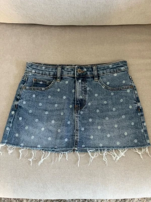 Blå jeanskjol med prickigt mönster - Säljer en blå jeanskjol med ljusa prickar och rå fransad kant nedtill. Kjolen har klassisk femficksdesign, bälteshällor och knappstängning framtill. Skriv för mått
