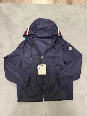 Mörkblå vindjacka från Moncler - Snygg mörkblå vindjacka från Moncler med huva och dragkedja framtill. Jackan har vita och röda detaljer på huvan samt Moncler-logga på ärmen. Tillverkad i ett glansigt, lätt material som står emot vind. Perfekt för dig som vill ha en stilren och sportig look.