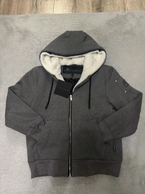 Grå hoodie från Moose Knuckles - Snygg grå hoodie från Moose Knuckles med dragkedja, stor huva och vit fluffig foder på insidan. Hoodien har svarta snören, två stora fickor fram och en logga på ärmen. Insidan är quiltad och ger extra värme. Perfekt för dig som vill ha både stil och komfort.