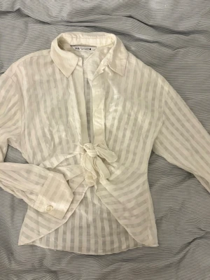 Vit randig skjorta med knyt från Zara - Säljer en vit skjorta från Zara i storlek XS med diskreta ränder och knyt framtill. Skjortan har klassisk krage, långa ärmar med knapp vid manschetten och är tillverkad i ett lätt och luftigt material som passar perfekt för en cool look.