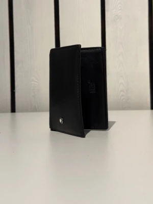 Montblanc Korthållare - Elegant svart läderplånbok från Montblanc i en klassisk och stilren design. (Vintage modell).                  Tillverkad i mjukt kvalitetsläder med flera kortfack och praktiskt ID-fönster. En perfekt plånbok för dig som vill ha något diskret men exklusivt i vardagen. Plånboken är använd och har några mindre skavmärken från normal användning. Den har nyligen behandlats med läderolja för att återfukta lädret och ge den bättre lyster och hållbarhet. Sömmar och funktion är i okej skick. 