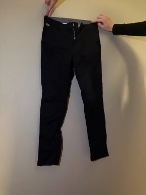 Mörkblå chinos från lacoste - Säljer ett par mörkblå chinos med klassisk femficksdesign och dragkedja. De har en snygg, smal passform och är tillverkade i ett stretchigt material som ger en bekväm känsla. 