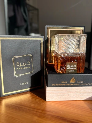 Lattafa Khamrah Eau de Parfum 100ml - Lattafa Khamrah Eau de Parfum kommer i en lyxig, fyrkantig glasflaska med detaljerat mönster och guldlabel. Doften har en varm bärnstensfärg och flaskan levereras i en elegant svart och guld ask. Perfekt för dig som gillar exklusiva parfymer med snygg design.