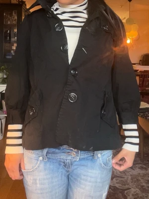Svart kort trenchcoat med stora knappar - Stilren svart trenchcoat med stora knappar framtill och fickor med lock. Perfekt till våren!.