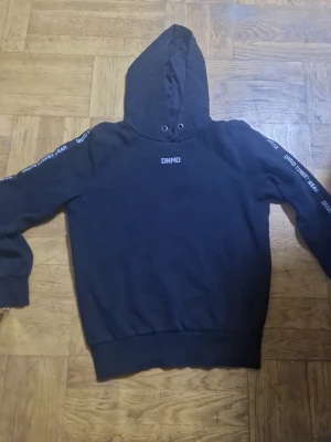 Svart hoodie från DNMD Streetwear - Svart hoodie från DNMD Streetwear med huva och ribbade muddar. Tryckt logga på bröstet och texten 'DNMD STREET WEAR' längs båda ärmarna. Klassisk passform och mjukt material, perfekt för en avslappnad stil.