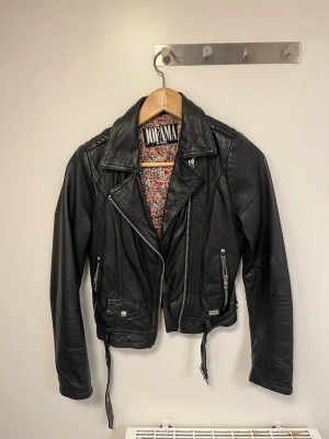 Svart skinnjacka från Jofama by Kenza - Snygg svart skinnjacka från Jofama by Kenza med coola detaljer som dragkedjor, axelvaddar och bälte i midjan. Insidan har ett blommigt foder som ger extra stilpoäng. Perfekt för dig som vill ha en klassisk bikerlook med en twist.