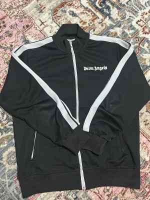 Svart ziptröja från Palm Angels - Snygg svart ziptröja från Palm Angels med vita ränder längs ärmarna och logga på bröstet. Tröjan har hög krage, hel dragkedja framtill och ribbade muddar. Perfekt för en sportig och trendig look.