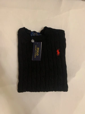 Svart kabelstickad tröja Polo Ralph Lauren - Svart kabelstickad tröja från Polo Ralph Lauren med klassisk rund hals och lång ärm. Tröjan har det ikoniska röda Polo-logot broderat på bröstet och ribbade muddar. Materialet är mjukt och skönt, perfekt för att hålla dig varm under kyliga dagar. Mått: längd: 60cm. Axelbredd: 35cm