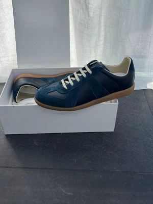 Blå sneakers från Maison Margiela Storlek 43 - Säljer ett par blå sneakers från Maison Margiela med klassisk design. Skorna har en kombination av mocka och läder, vita snören och en ljusbrun gummisula. Perfekt för dig som gillar stilrena och trendiga sneakers med premiumkänsla. Kvitto kan skickas! Storlek 43 och pris kan diskuteras . 🤙