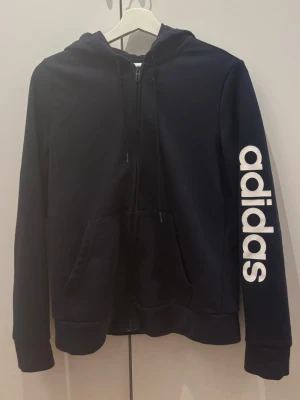 Svart hoodie från Adidas med tryck - Snygg svart hoodie från Adidas med stor vit logga längs ena ärmen. Klassisk modell med huva, dragsko och känguruficka framtill. Perfekt för en sportig och avslappnad stil.