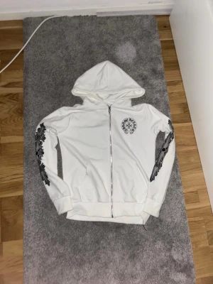 Vit Chrome Hearts zip hoodie - Vit hoodie med dragkedja från Chrome Hearts. Har svarta tryck längs båda ärmarna och en rund logga på bröstet. Utrustad med huva och två fickor framtill. Materialet är mjukt och bekvämt, perfekt för en avslappnad stil. Band finns till och kan passa till M också.