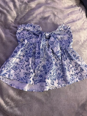 Blommig vit och blå blus med knyt - Supersöt blommig blus i vitt och blått med volangdetaljer och knyt framtill. Toppen har en smockad rygg och är ärmlös med rynkade axelband. Perfekt för en fräsch och somrig look.