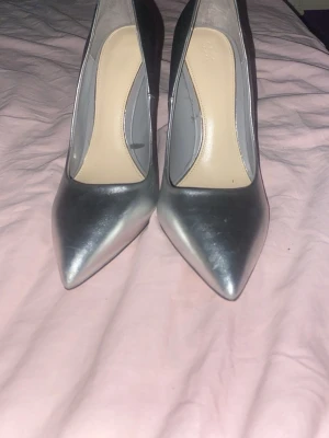 Silvriga spetsiga pumps med hög klack - Snygga pumps i metallic-silver med spetsig tå och hög smal klack. Skorna har en stilren design och är tillverkade i ett glansigt material som ger en modern look. Perfekta för dig som vill sticka ut med ett par trendiga klackar.1199