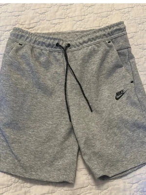 Grå sweatshorts från Nike - Snygga grå sweatshorts från Nike med svart snörning i midjan och diskret logga på benet. Shortsen har två sidofickor och är tillverkade i mjuk bomullsmix som passar perfekt till avslappnade dagar.