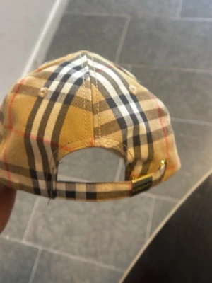 Burberry keps med klassiskt rutmönster - Snygg keps från Burberry med det ikoniska beigea, svarta och röda rutmönstret. Justerbar rem baktill med metallspänne och märkets logga på insidan. Perfekt accessoar för att lyfta din stil med en lyxig touch.