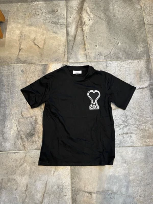 Svart t-shirt från Ami med hjärtdetalj - Snygg svart t-shirt från Ami med ett stort vitt hjärta och bokstaven A broderat på bröstet. Klassisk rund halsringning och korta ärmar. Tillverkad i mjuk bomull som känns skön mot huden. Perfekt för en clean och stilren look.