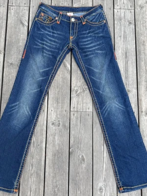 Vintage True Religion low rise jeans blå  - Blå bootcut jeans från True Religion med orange kontrastsömmar och broderade bakfickor. Jeansen har slitningar framtill och klassisk femficksdesign. Tillverkade i denim med snygg tvätt och detaljerade sömmar för en cool look. Skriv privat om fler bilder/detaljer önskas 